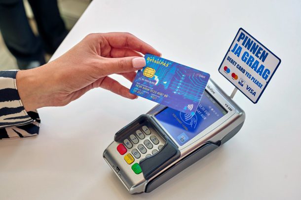 Hand houdt nieuwe Debit betaalpas boven betaalautomaat met 'PINNEN JA GRAAG' bordje, symboliseert acceptatie van nieuwe passen.