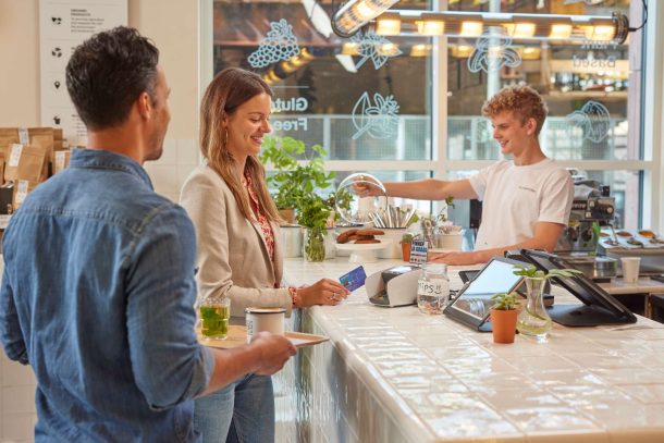 Jonge vrouw betaalt met betaalpas bij betaalautomaat aan de counter van een horecazaak. Naast haar staat een man met dienblad met daarop thee en koffie.