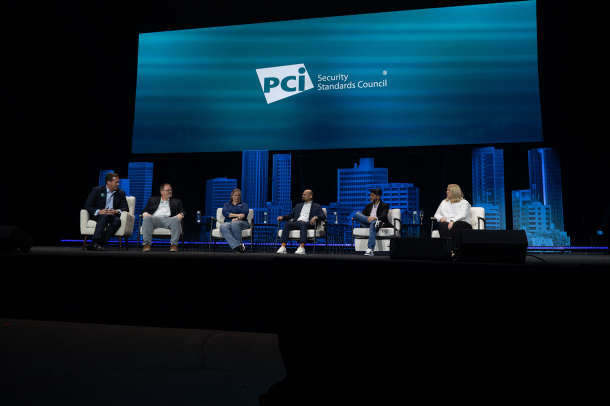 4 mannen en 2 vrouwen zitten op witte stoelen op een podium staan. Op de achtergrond in het blauw de skyline van een stad en daarboven heel groot op een scherm het logo van PCI SSC.