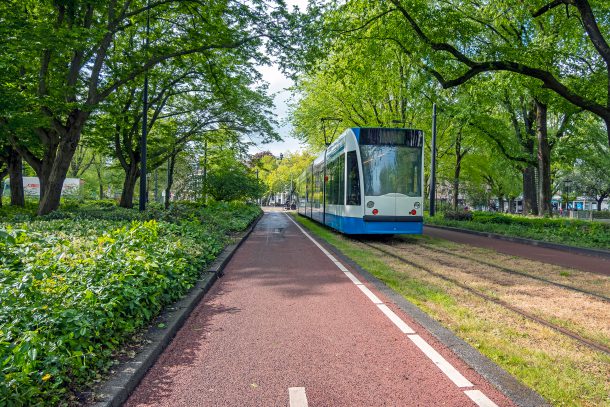 Vanaf een fiets genomen, groene Amsterdamse omgeving met centraal een GVB-tram die voorbijrijdt.